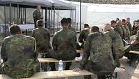 Zahlreiche Soldaten in Uniform sitzen auf Bänken, ein Militärpfarrer steht vor ihnen und predigt.