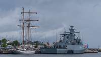 Ein graues Schiff liegt vor einen weißem Segelschiff. Beide haben an einer Pier festgemacht.