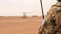 Ein weißer UN Hubschrauber startet. Ein Soldat mit einem blauen Ärmelabzeichen beobachtet den Vorgang