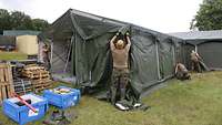 Soldaten bauen ein Bundeswehrzelt auf. Davor stehen Kisten auf der Wiese.