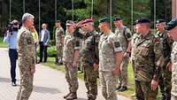 Mehrere Soldaten verschiedene Nationen stehen bei einer Zeremonie auf einem betonierten Platz vor vielen Flaggenmasten.