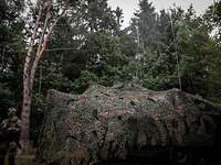 Soldat auf einer Waldlichtung steht neben einem mit Tarnnetz überzogenen Lkw, auf dem zwei Antennen aufgebaut sind.