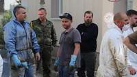 Mehrere Männer, zum Teil in Uniform oder in Schutzanzügen, stehen im Freien