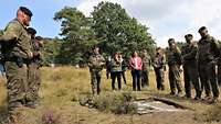 Inmitten vieler Soldaten im Flecktarnanzug steht eine Zivilistin bei einer Ausbildung im Gelände auf einer grünen Wiese.