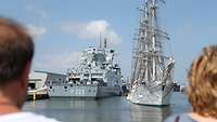 Ein graues Schiff liegt an Pier und wird von einem weißem Segelschiff passiert.