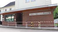 Ein flaches Backsteingebäude mit großem Fenster, an der Wand ein metallenes Schild mit der Schrift „Oberst-Herrmann-Kaserne“