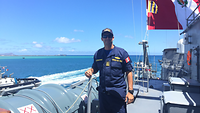 Der Marineoffizier aus Peru steht an Bord eines Schiffes. Daneben ist das Meer zu sehen, hinter ihm weht die peruanische Flagge