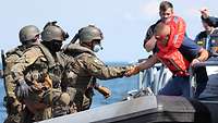 Drei Personen in Flecktarn-Uniform auf einem Gummiboot übernehmen eine Person in Schwimmweste an Bord.
