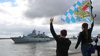 A grey warship in the water of a harbour; in the foreground on a pier two waving people.