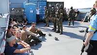 Mehrere Gerettete sitzen an Deck des Schiffes und werden durch Soldaten in Uniform bewacht.