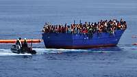 Ein blaues Boot mit vielen dunkhäutigen Menschen treibt auf offener See.