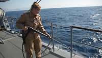 Eine Soldatin mit einem Hochdruckreiniger in der Hand, steht an der Reling eines Schiffes und spritzt sie ab