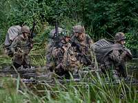 Vier Soldaten mit schwerem Gepäck und Waffen im Anschlag waten durch einen Fluss.