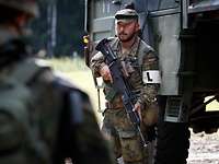 Dicht vor einem Fahrzeug steht ein Soldat mit der Waffe im Anschlag.