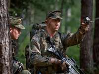 Ein Soldat hält einen Kompass und streckt seinen Arm damit auf Schulterhöhe aus und kneift ein Auge zu.