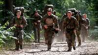 Auf einem staubigen Waldweg laufen mehrere Soldaten. Auf dem Rücken tragen sie ihre großen, vollgepackten Rucksäcke.