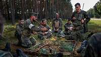 Am Waldrand hocken viele Soldaten um einen Geländesandkasten herum. Der Ausbilder erklärt das spätere Vorgehen.