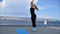 Eine Soldatin in Sportkleidung springt vor ihrer blauen Turnmatte in die Luft. Sie macht Sport auf dem Flugdeck eines Schiffs