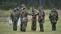 Mehrere Soldaten stehen auf einem grünen Feld. Zwei von ihnen legen ihre Hand auf eine gesenkte Fahne. 