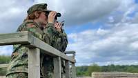 Soldaten und Soldatinnen in Uniform, die mittels eines Fernglases in das Gelände blicken und dieses damit erkunden.