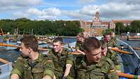 Eight soldiers in battle dress in a rowing boat on the water with a large red building in the background.