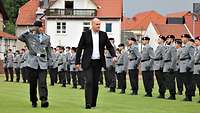 Ein Zivilist und ein grüßender Soldat schreiten eine Front von Soldaten auf einer Wiese ab.