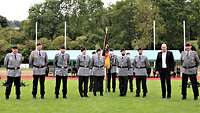 Soldaten in grauer Uniform stehen neben einem Zivilisten auf einer Wiese mit der Truppenfahne.