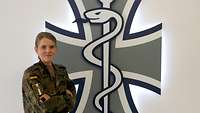 Oberleutnant Elisabeth Motzka im Portrait Eine Soldatin steht vor einem beleuchteten Logo: Äskulapstab vor dem Eisernen Kreuz der Bundeswehr