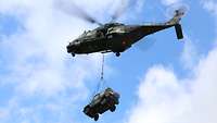 Ein Hubschrauber fliegt unter blauem Himmel. Er transportiert an einem Stahlseil einen Geländewagen.