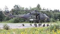 Acht Soldaten auf einer grünen Wiese sichern kniend die Umgebung. Der Hubschrauber schwebt über ihnen. 