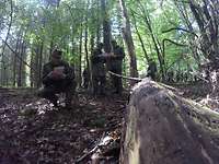 Ein Gefreiter hockt auf dem Waldboden, um mögliche Kampfmittel zu identifizieren. Kameraden notieren ihre Beobachtungen.