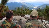 Ein belgischer und ein deutscher Soldat hocken hinter einem Felsen mit Blick auf die Alpen.