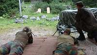 Zwei belgische Soldaten liegen auf dem Boden mit jeweils einem Maschinengewehr. Ein deutscher Soldat erklärt die Besonderheiten.