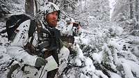 Zwei Soldaten knien mit ihrer Ausrüstung im verschneiten Wald, einer schaut durch ein Fernglas in die Ferne