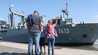 Ein Mann und 2 Kinder blicken auf ein graues Schiff.