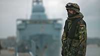 Ein Soldat in Flecktarnuniform und mit Stahlhelm steht in einem Hafen vor einem grauen Schiff.