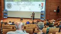 Colonel Frank Wasgindt stands in front of a large screen and speaks to the audience