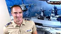 A soldier is smiling and standing in front of a wall with a poster that shows two ships and an aircraft