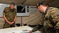 Ein Marineoffizier in Flecktarn lässt sich vor einem Tisch von einem anderen Soldaten etwas erklären.