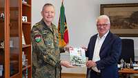 Ein Soldat und ein Zivilist in dunkelblauem Anzug zeigen in einem Büro gemeinsam ein Magazin in die Kamera.