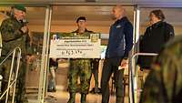 Ein Soldat hält einen symbolischen Spendencheck hoch, daneben stehen weitere Personen.