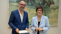 Ministerin Kramp-Karrenbauer und eine männliche Person, die ein Dokument in der Hand hält, stehen lächelnd nebeneinander