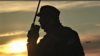 Ein Soldat spricht draußen bei Sonnenuntergang in ein Funkgerät. Am goldenen Himmel fliegt ein kleines Flugzeug.