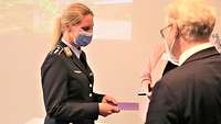 Ein Mann in schwarzem Anzug überreicht einer Soldatin in blauer Uniform ein Kästchen und eine Urkunde.