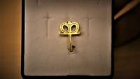 In einer geöffneten Schmuckschatulle liegt mittig ein goldenes Kronenkreuz, ein Pfahl mit einer einfachen Krone.