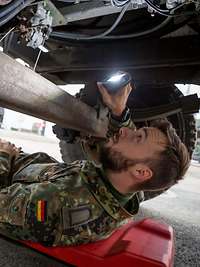 Ein Soldat liegt unter einem Fahrzeug und leuchtet mit einer Taschenlampe in das Fahrwerk