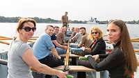 11 Männer und Frauen sitzen in einem Ruderboot und schauen in die Kamera