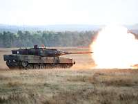 Ein Kampfpanzer steht auf einer großen braunen Wiese. Ein heller Feuerball verlässt die Kanone.