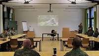 Mehrere Soldaten sitzen an Tischen, die in U-Form vor einer Projektionsfläche aufgebaut sind.