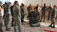 Mehrere Soldaten stehen in einem Stall auf sandigem Boden um die Ausrüstung eines Tragtieres herum.